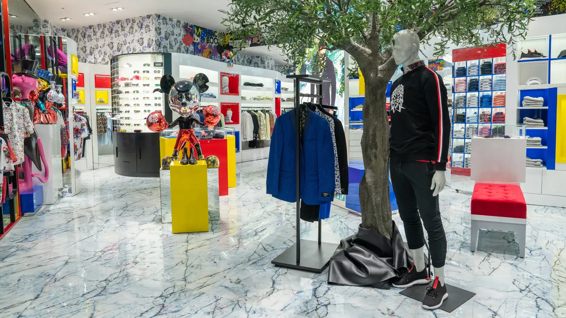 Menswear boutique interior with polished marble floors, an indoor olive tree and a mannequin in a black and red track jacket beside a chrome cartoon-mouse sculpture