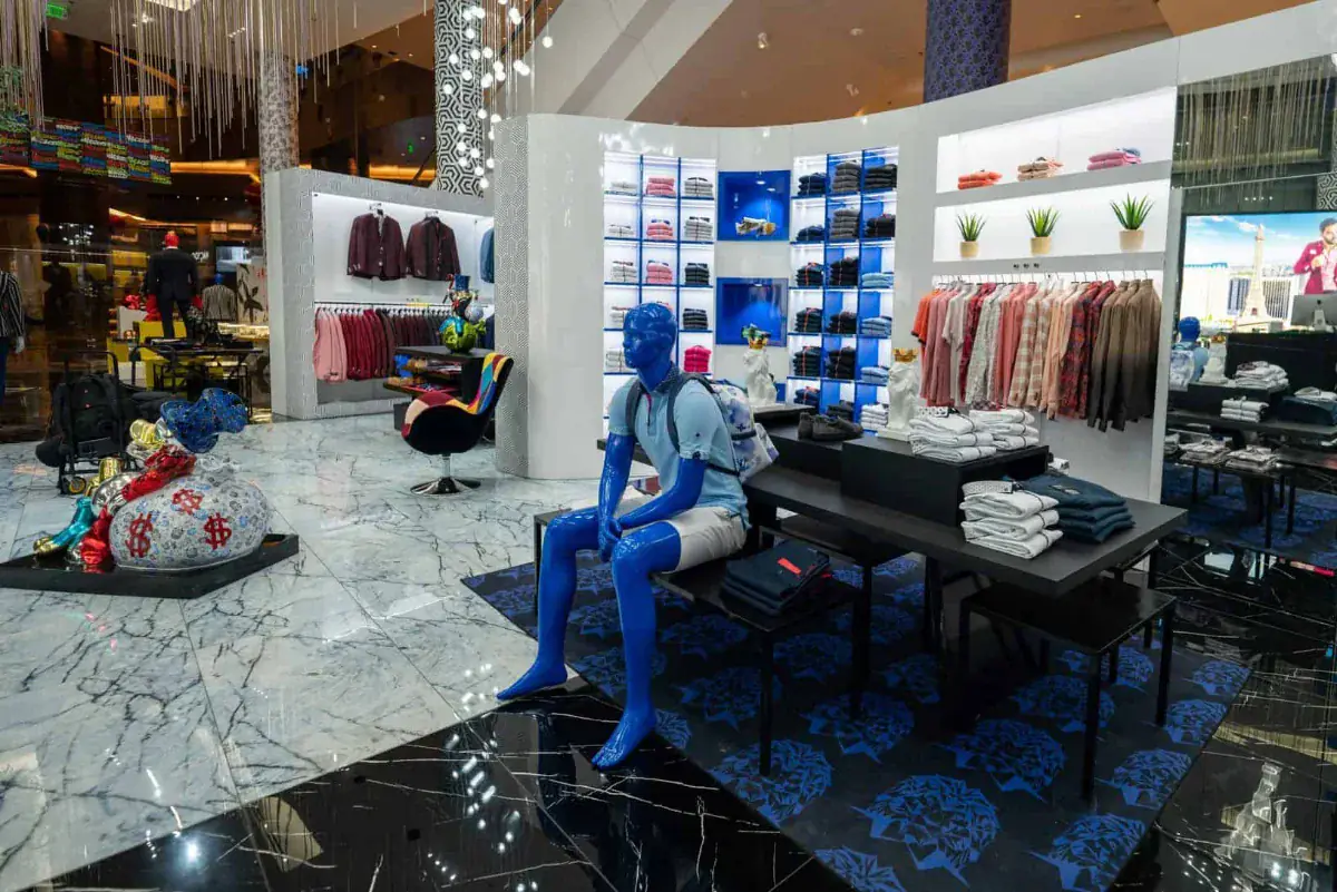 Blue-lacquered seated figure beside a black display table stacked with folded shirts, backlit cubby shelving of knitwear behind