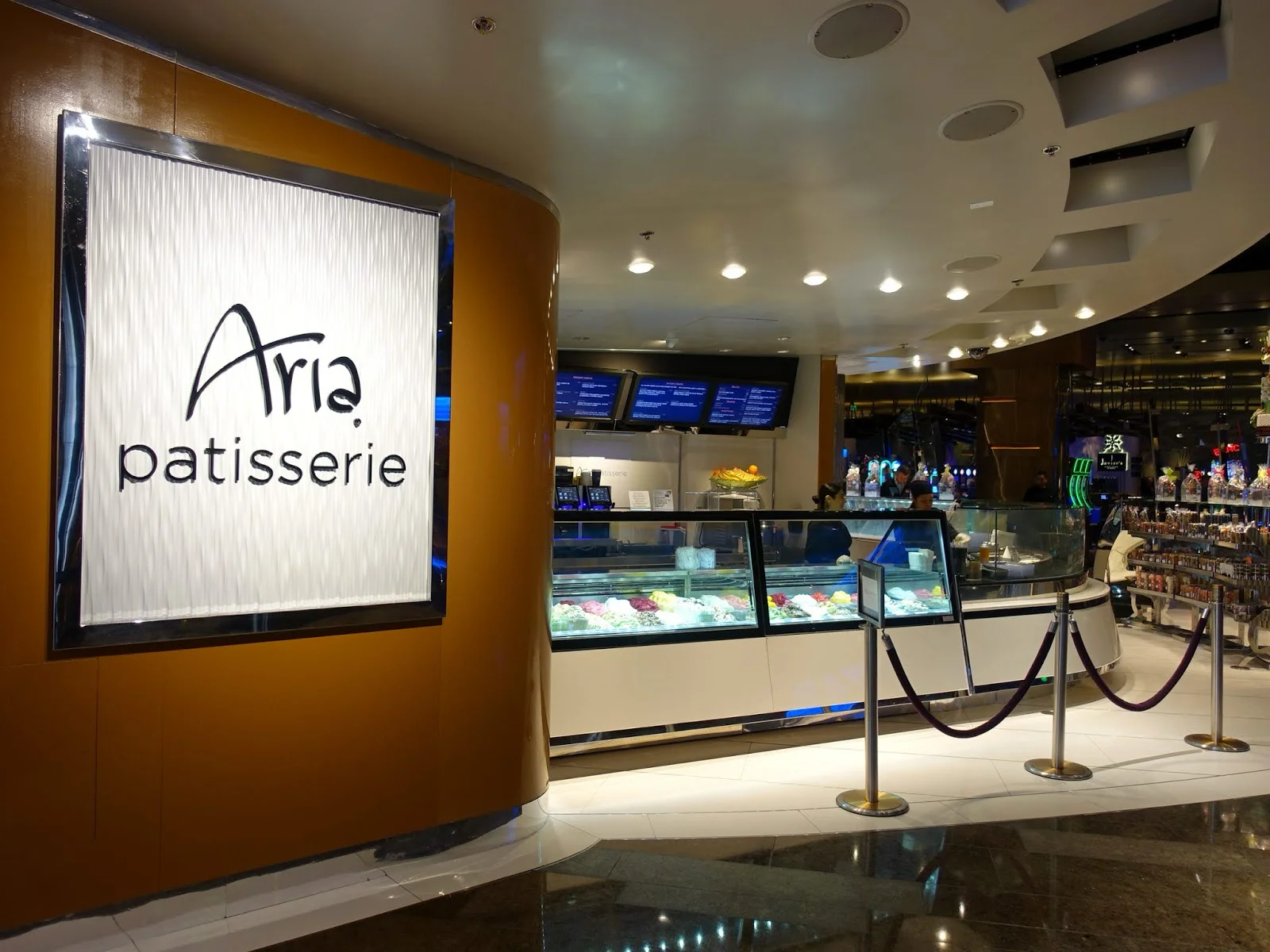 Backlit Aria Patisserie sign on a curved caramel-wood wall beside an illuminated gelato and pastry display case roped off with velvet stanchions inside ARIA Resort