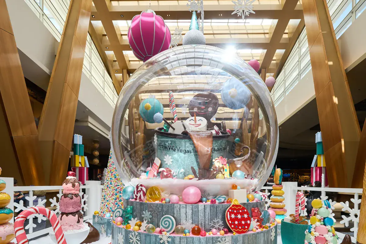Holiday candyland display at ARIA with a giant snow globe holding a gingerbread man and snowman in an oversized coffee cup, surrounded by oversized ornaments, candy canes, and macaron towers