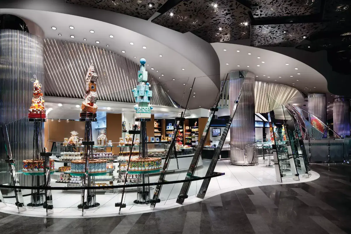 Sculptural ARIA Patisserie interior with fluted silver columns, an undulating white ceiling under a patterned black canopy, and glass tables of packaged sweets beneath whimsical tiered cake sculptures
