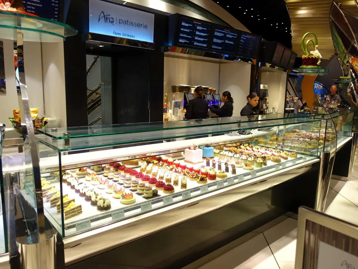 Aria Patisserie counter marked Open 24 Hours with a long glass case displaying rows of individual French pastries, fruit tarts, and cakes as staff work behind it