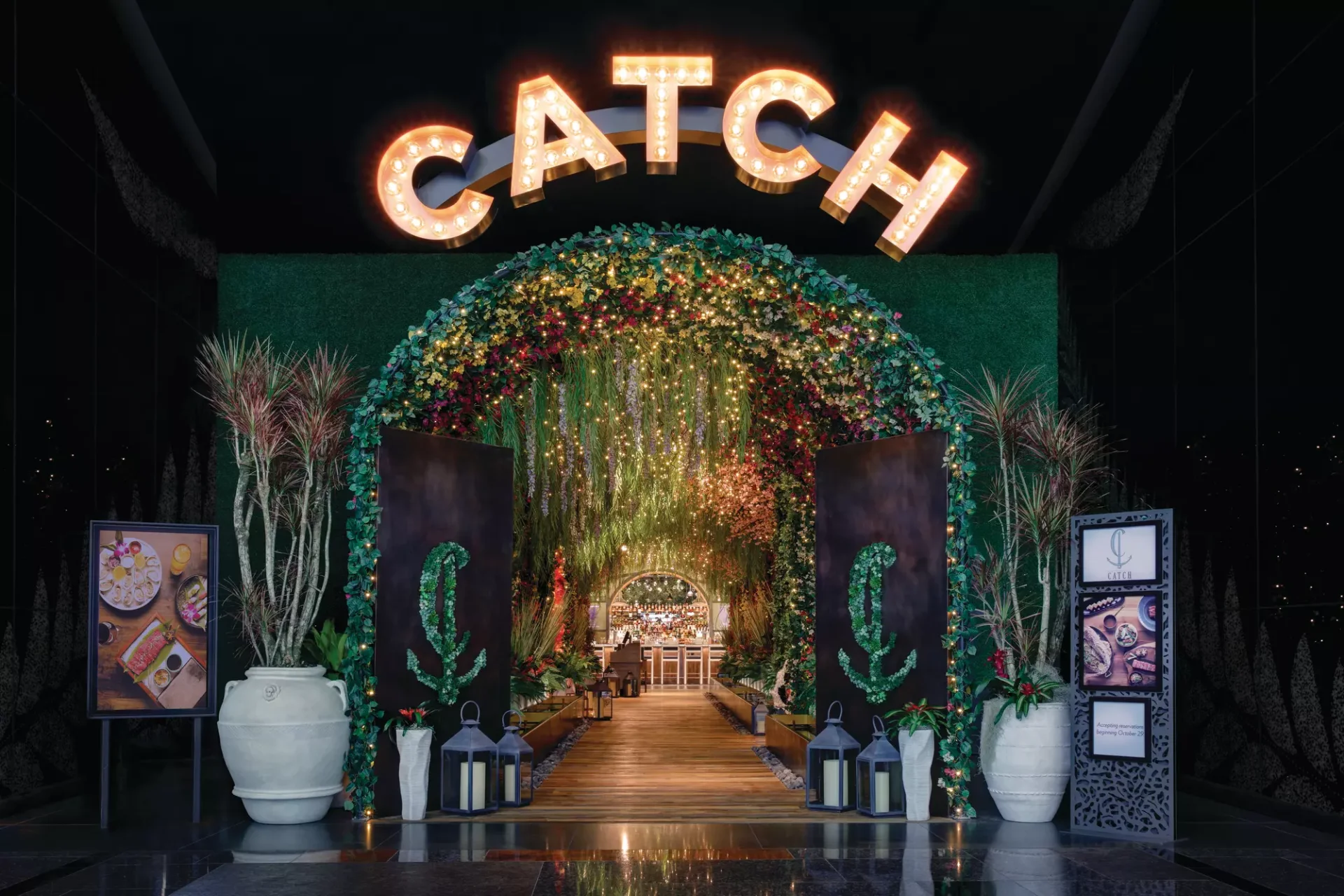 Illuminated CATCH marquee sign above a floral archway entrance with anchor logos, lanterns, and a wooden walkway leading to the bar at CATCH Las Vegas in ARIA