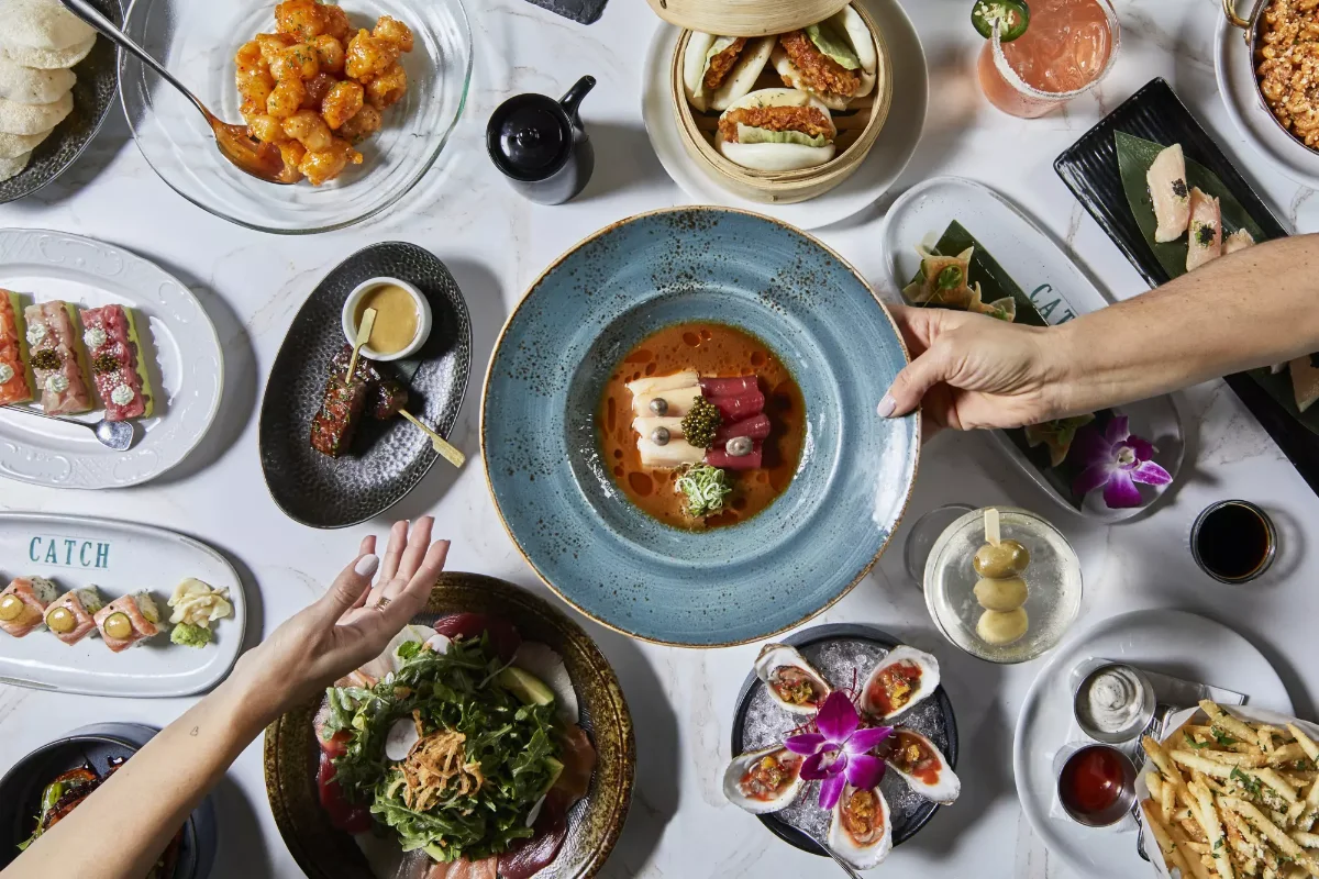 Overhead spread of CATCH Las Vegas dishes including crispy shrimp, bao buns, sashimi rolls, oysters on ice, truffle fries, and cocktails on a marble table