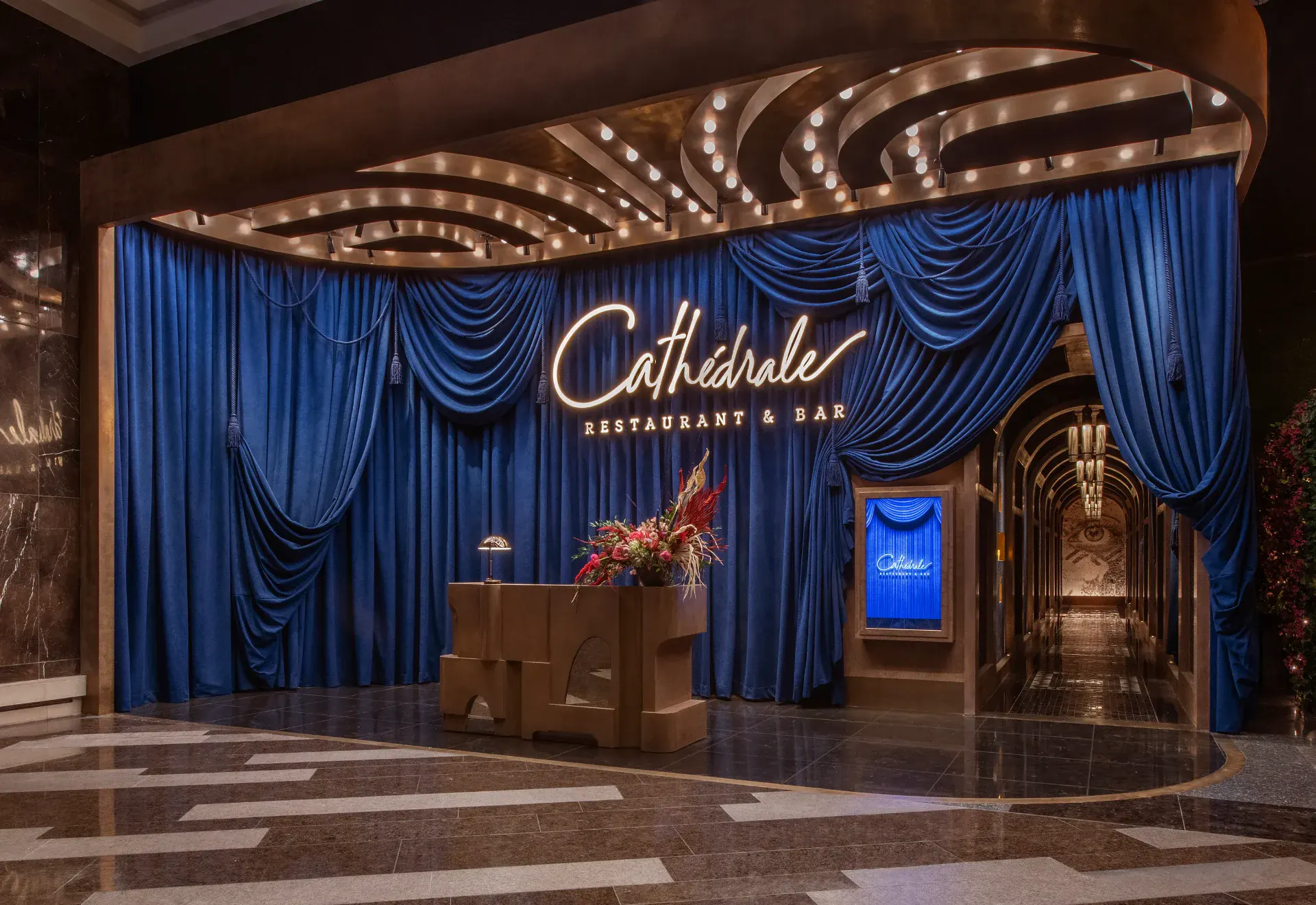 Illuminated script Cathédrale Restaurant & Bar neon sign above deep-blue velvet draperies at the ARIA entrance, with a stone host stand, a tall floral arrangement, and an arched mirrored corridor leading into the dining room