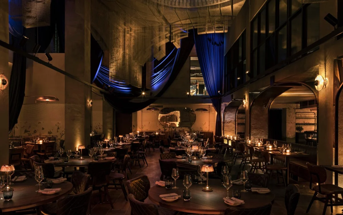 Soaring double-height Cathédrale dining room lit by candlelight, with draped blue and woven-mesh fabric suspended from the ceiling and a sculptural illuminated wall feature
