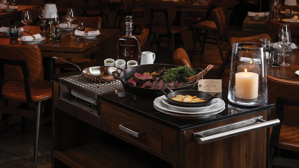 Tableside service cart at Cathédrale with a cast-iron pan of sliced rare côte de boeuf and herbs over a grill, a skillet of sliced potatoes, a Woodford Reserve bottle, and a candle in a hurricane glass