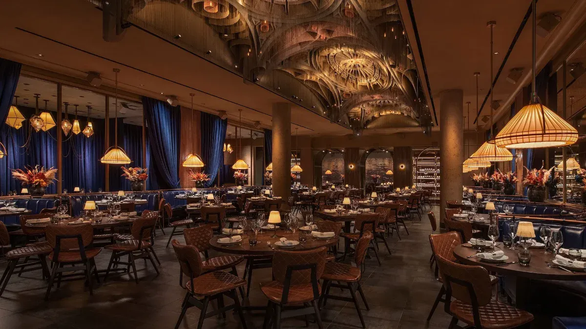 Wide Cathédrale dining room beneath the dramatic cathedral-inspired sculptural ceiling, with woven pendant lamps, blue velvet curtains, leather chairs, and dried-floral centerpieces