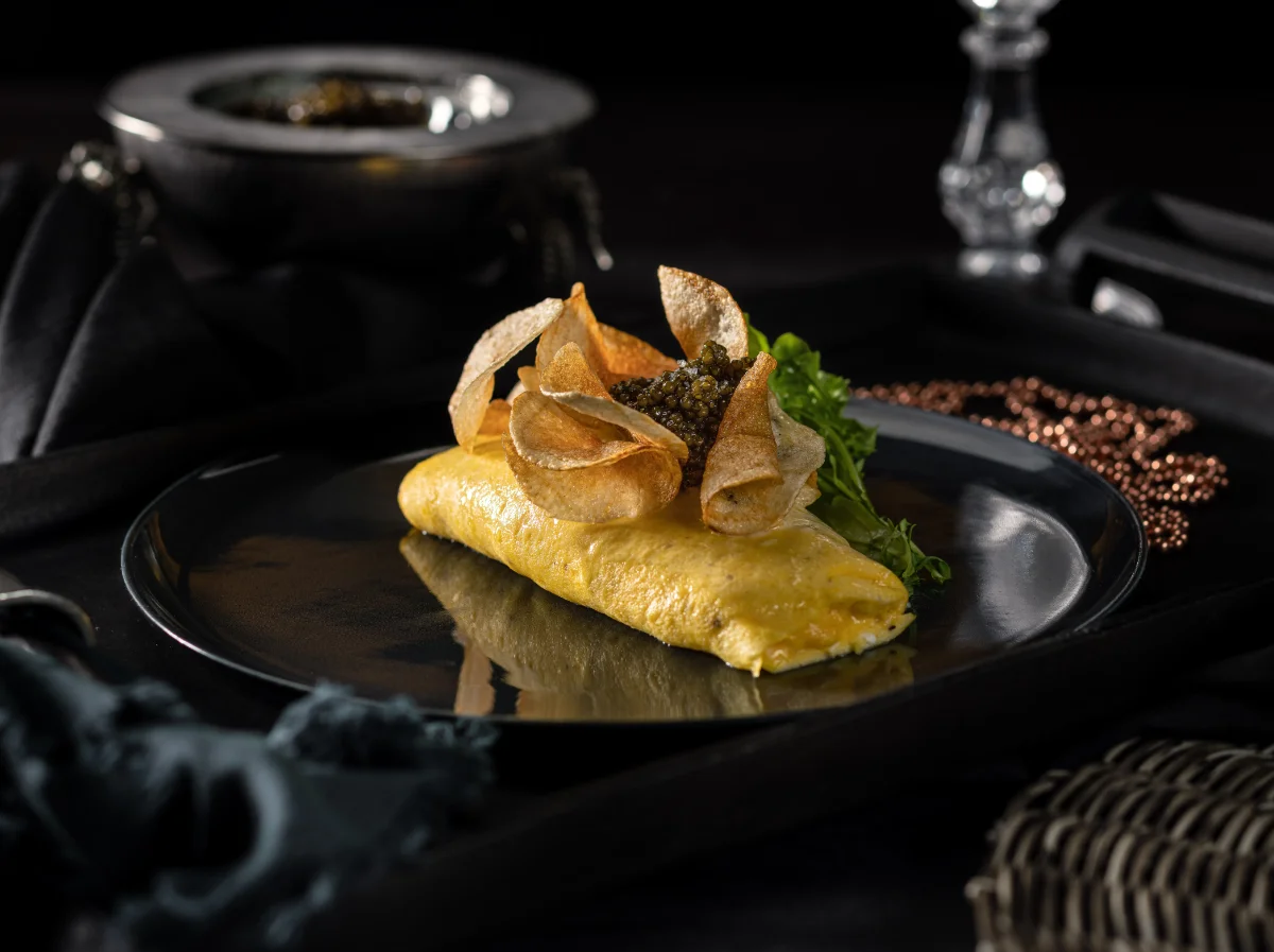 Folded omelette topped with caviar and crisp potato chips and a sprig of greens on a black plate in Cathédrale's dark, moody table setting