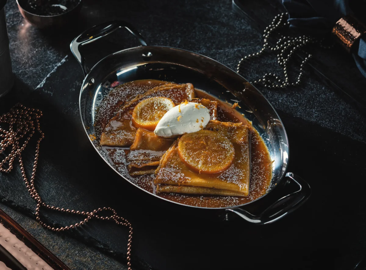 Crêpes Suzette dessert in a silver pan with caramelized orange slices, powdered sugar, and a quenelle of cream on a dark marble table