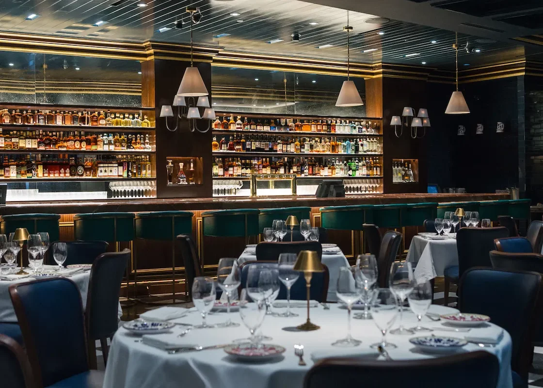 Din Tai Fung dining room with a long backlit bar of liquor bottles, green chairs, and white-clothed tables set with wine glasses