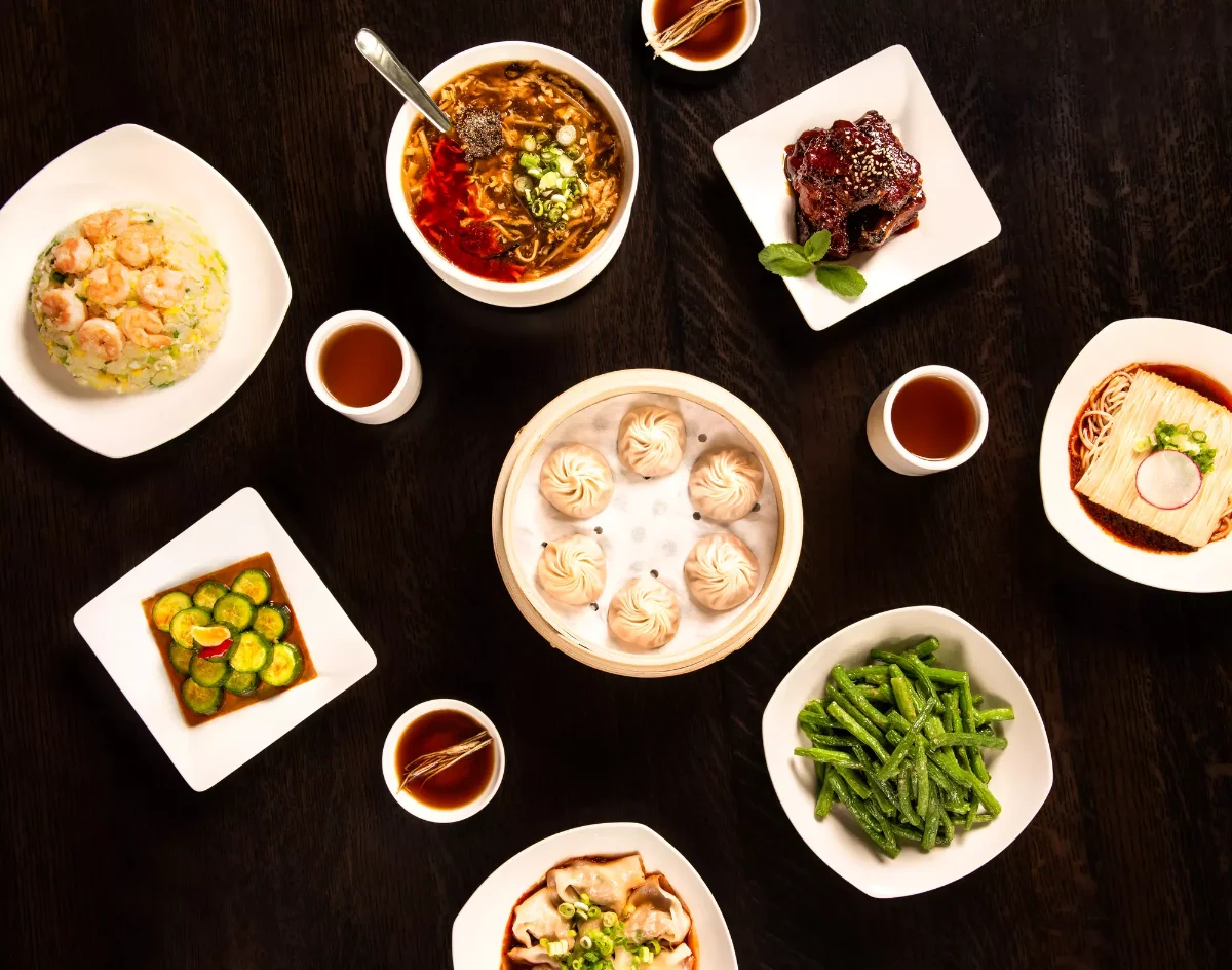 Overhead spread of Din Tai Fung dishes including a bamboo steamer of xiao long bao soup dumplings, shrimp fried rice, hot and sour soup, green beans, cucumber salad, and tea
