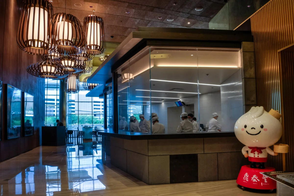 Glass-walled open kitchen with chefs in white folding dumplings beside bamboo-cage lanterns and the Din Tai Fung dumpling mascot at ARIA