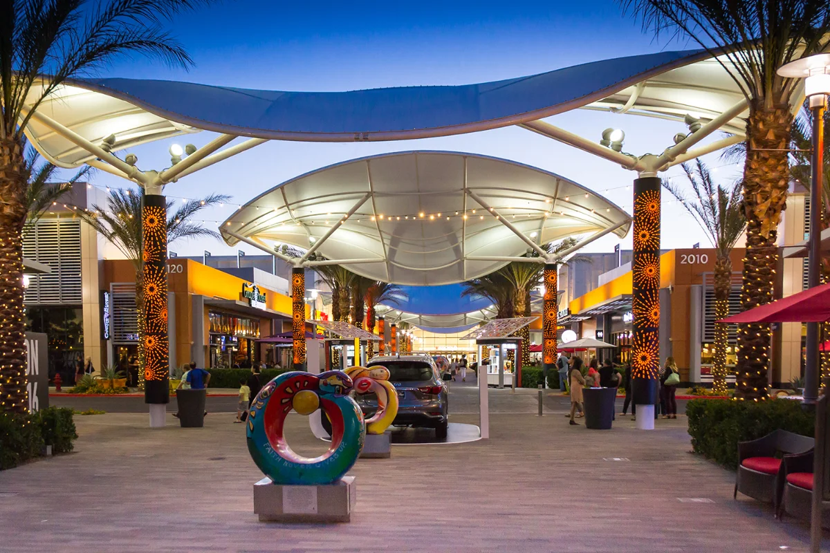 White sail shade canopies and palm trees lit at dusk above a colorful circular sculpture at Downtown Summerlin