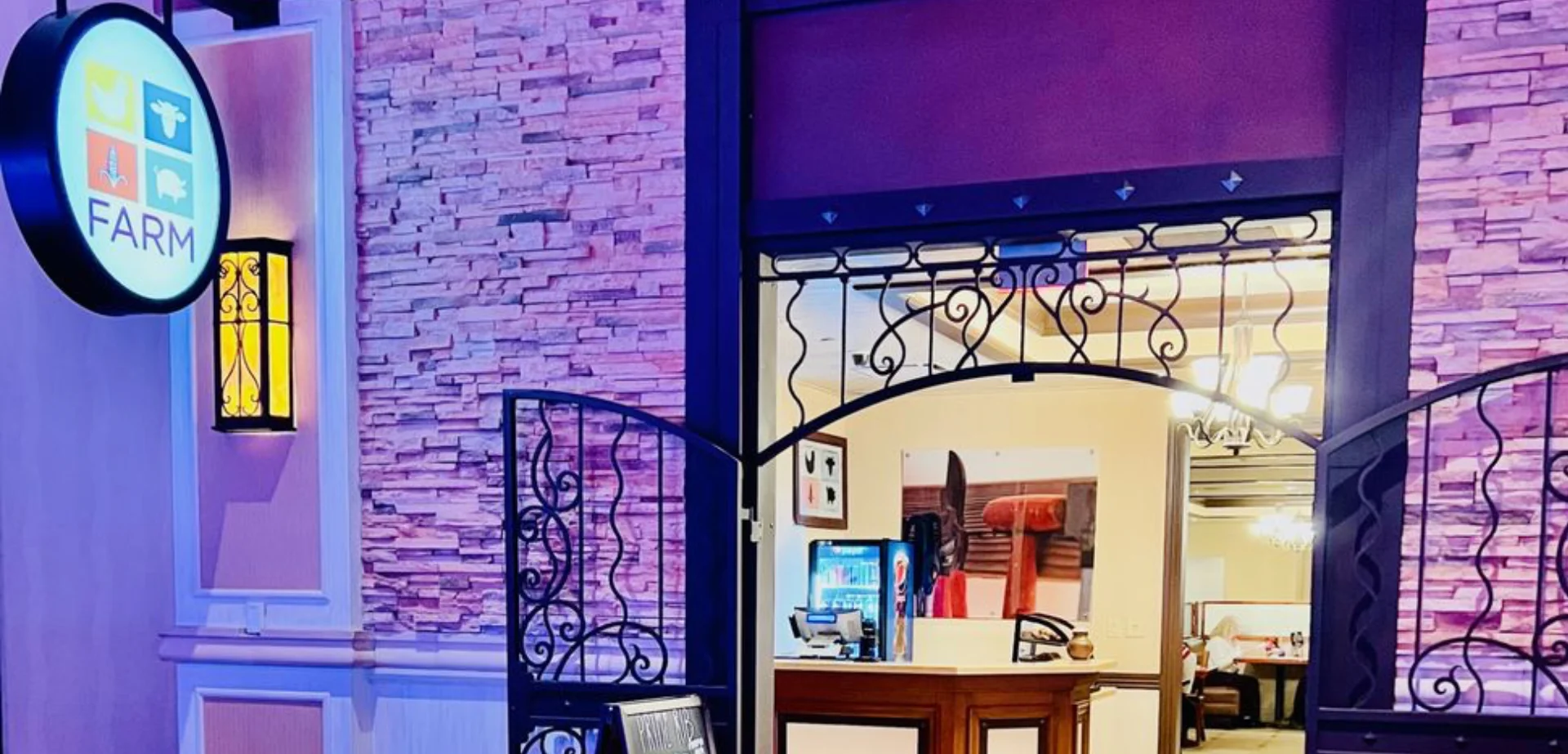 Illuminated round FARM logo sign beside a wrought-iron gate entrance with stacked-stone walls leading into the dining room at Aliante