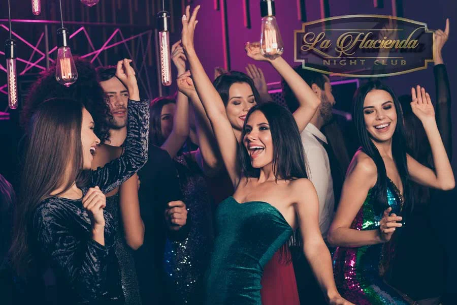 Group of partygoers in sequined and velvet cocktail dresses cheering with raised arms under Edison-bulb pendant lights on the nightclub dance floor