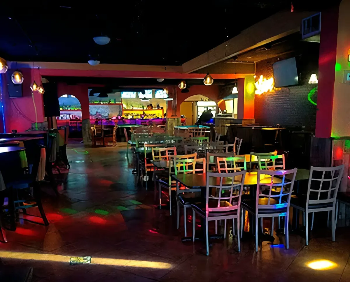 Empty club interior with clustered dining tables and chairs, arched doorways and a back bar washed in red, green and purple lighting