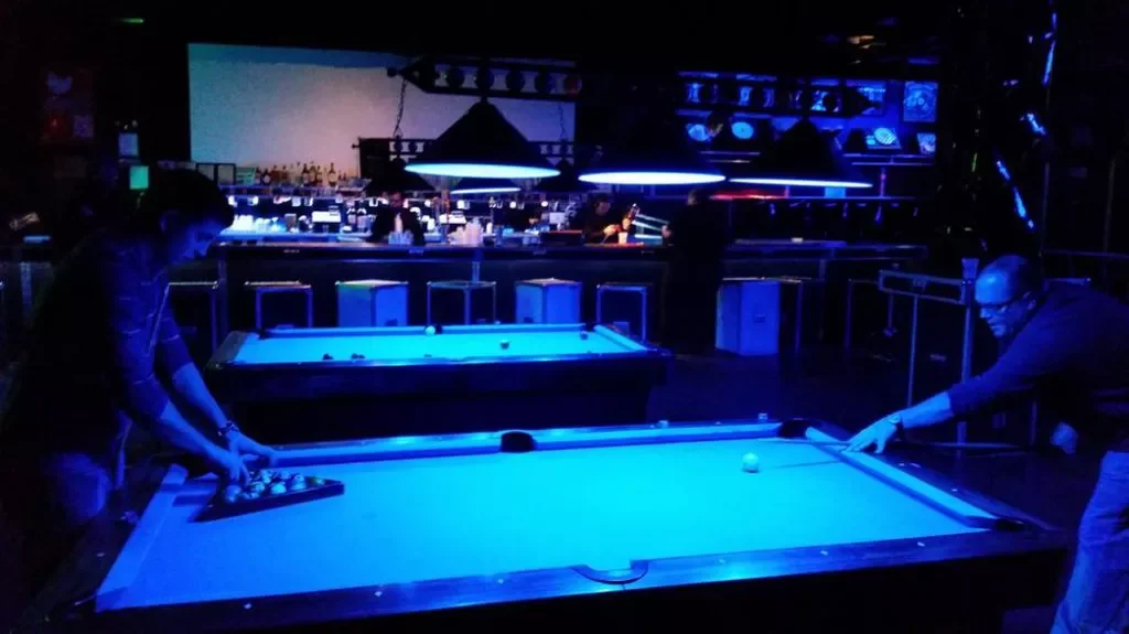 Two guests playing pool on felt billiards tables lit by blue neon beside the illuminated bar counter and stools