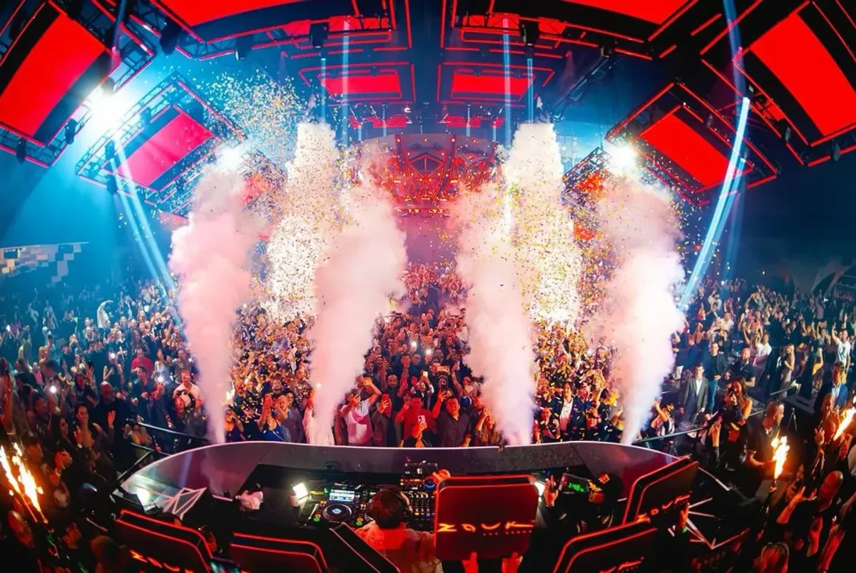 CO2 jets and gold confetti bursting over a cheering crowd facing a red-lit LED stage and elevated DJ booth during a high-energy set
