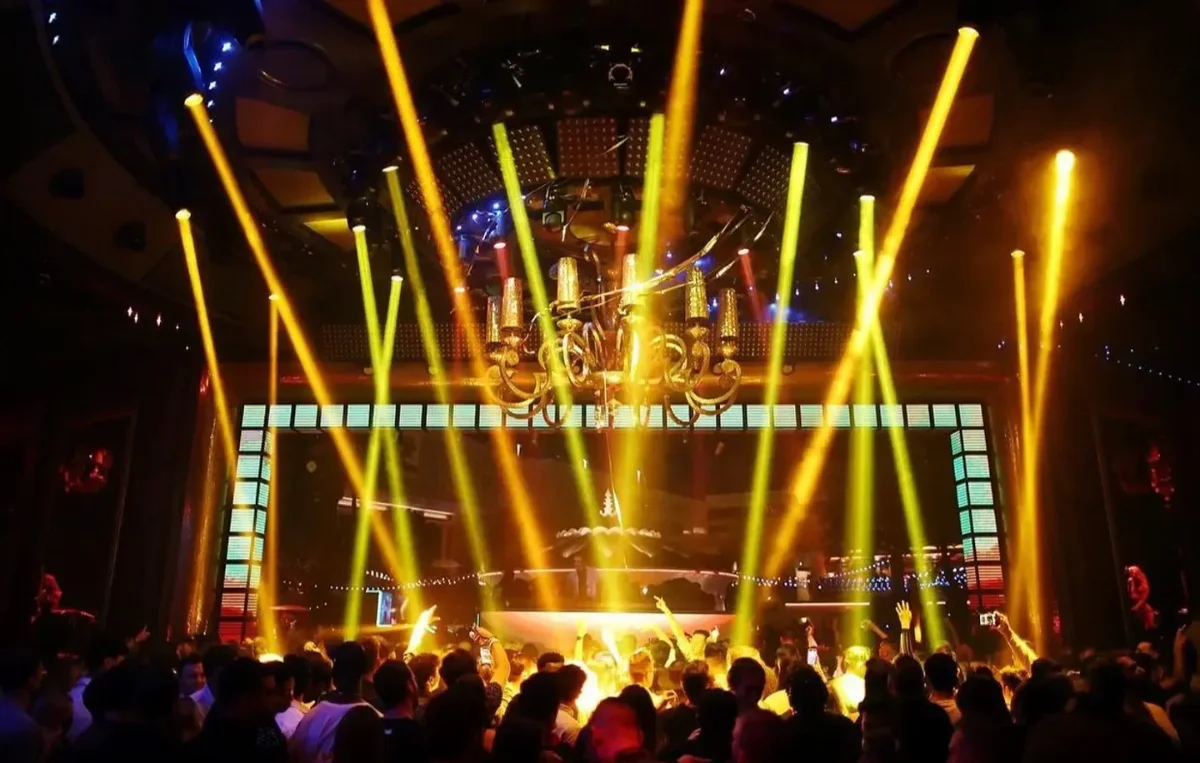 Yellow beam spotlights fanning out from an ornate gold chandelier over a crowd with raised arms in front of a glowing LED backdrop
