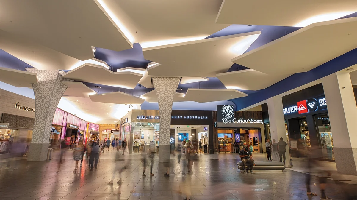 Shoppers crossing a wide concourse under angular illuminated ceiling panels past storefronts at Miracle Mile Shops